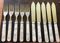 Photo showing six knives with pale yellow handles next to six forks with mother of pearl handles, presenting the complete set of 12 vintage cutlery items.