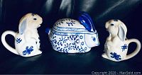 Front view of two rabbit-shaped creamers and covered rabbit bowl showing white porcelain with blue painted details.