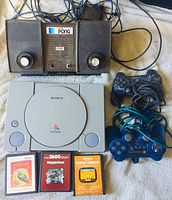 Top-down wide photo showing Atari Pong console, PlayStation 1 console with two controllers, and three Atari 2600 game cartridges.