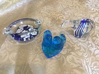 Lot of three glass art pieces on beige patterned fabric: oval ashtray, blue heart-shaped paperweight, and bird figurine