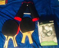 Four ping pong paddles, collapsible net, and a box of table tennis balls, laid out on a blue surface.