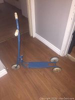 Blue vintage metal scooter standing indoors on a wooden floor showing full structure including grips and wheels