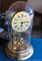 Front view of a gold tone Eurena anniversary clock under a clear glass dome showing a white face with Roman numerals and ornate black hands.