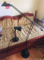 Two lamps displayed on a beige and red sofa—one tall black metal adjustable halogen floor lamp with rectangular shade, and one smaller black metal gooseneck desk lamp with round base and cone-shaped shade.