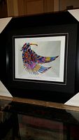 Framed and matted print of colorful Native American figure with wings, displayed in black frame with white matting corners visible.