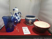Lot consisting of a blue and white ceramic teapot, a blue glass Shirley Temple pitcher, a blue jasperware style planter, and two ceramic bowls on a red surface.