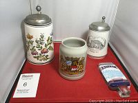 Three beer steins and one beer tap handle arranged on a red cloth background.