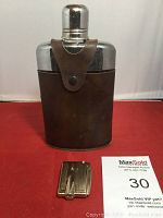 Metal flask with brown leather case and metal snap closure, approx. 8 inches tall, standing upright on a red surface with a MaxSold auction card visible.