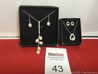 Two pearl style necklace and earring sets displayed in jewelry boxes on red surface.