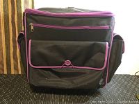 Front view of black crafting rolling case with a large flap pocket with button closure and purple trim