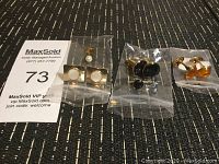 Four pairs of cuff links in clear plastic bags on a dark woven fabric surface, showing various styles and colors.