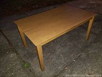 Side angled view of the wood rectangular dining table outside on pavement showing the table's light wood color and straight legs.