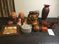 Photo showing all items on black table including vase, jars, bear figurine, feet figurines, spherical ceramic pieces, bird figurine, framed tile, coasters, and wooden box.