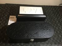 Black elongated jewelry case with embossed floral pattern and word 'fountainque' on lid, displayed closed on table.