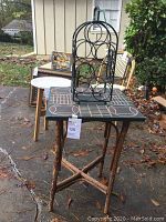 Rustic wooden side table with branch legs and painted tabletop with chair silhouette pattern, displayed outdoors with a metal wine bottle holder on top.