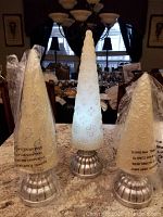 Three cone-shaped light up decorative trees on silver round bases, placed on a marble countertop in a dining room. Two trees wrapped in clear protective plastic packaging.