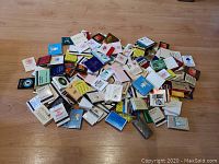 Wide shot of approximately 100 vintage matchbooks spread out on a wooden surface, showing a variety of advertising logos and colors.