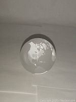 Front view of the clear glass paperweight showing the North American and South American continents etched in white.