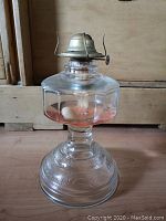 Full view of the glass oil lamp showing the faceted body, rounded embossed base, and metal burner.