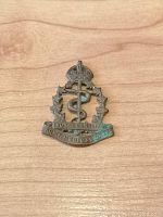 Front view of Royal Canadian Army Medical Corps cap badge showing crown, wreath, snake and staff insignia, along with inscription on banner.