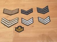 Seven assorted vintage embroidered military patches laid on a wooden surface, showing chevron stripes and a maple leaf emblem patch.