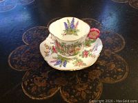 Cup and saucer displayed on dark patterned surface showing floral patterns and flower-shaped handle
