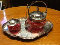 Overall view of two cranberry glass jam jars with silver tops and spoon on an English plated gallery tray on wooden table