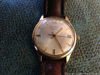 Front view of the wristwatch showing cream dial, gold-tone markers and hands, date window, and small crack in crystal.