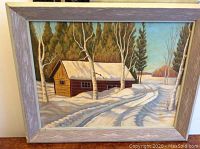Framed oil painting of snow-covered cabin surrounded by trees with visible snow tire tracks and shadowed ground.