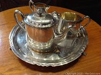 Four-piece silver-plated teaset on a wooden table showing tray, teapot, sugar bowl, and creamer.