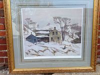 Framed print showing winter landscape with houses in snow, framed in a gold ornate frame.