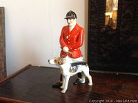 Side view of the porcelain figurine showing the master of hounds in a red jacket and black helmet with the hunting dog next to him.