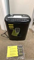 Front view of black Amazon Basics paper shredder with crack visible on the bottom paper bin, placed on carpeted floor with power cable attached.