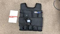 Front view of black Runmax weighted vest lying flat, showing front panel, straps, and manual beside it.