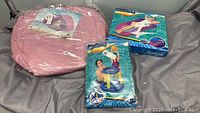 Three packaged inflatable floaters laid on a gray surface: pink circular floater, blue circular floater, and Bestway basketball hoop float with net and legs.