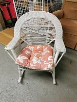 Front view of the white painted wicker rocking chair with reddish-orange floral cushion on the seat, showing the woven wicker backrest, armrests, and curved rocking legs.