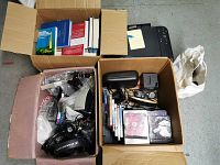 Top-down photo showing two open cardboard boxes containing books, electronics, headphones and office supplies, printer in background.