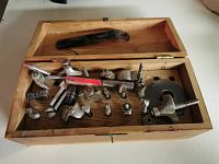 Wooden box filled with various specialty drill bits and metal accessories for woodworking and drilling tasks.