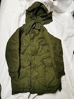 Full view of the olive green military parka laid flat showing front buttons, hood, and large front pockets.