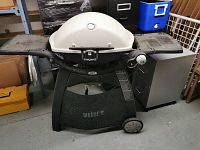 Front view of beige Weber Q grill on black wheeled stand with side tables