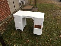 Full front view of white painted solid wood antique desk/vanity on grass outside. Shows 4 drawer doors, one open, and recessed seating area.