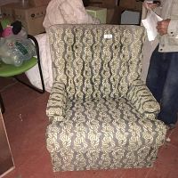Photo of an upholstered chair with a leaf-patterned fabric in light and medium brown tones, showing a tufted backrest and armrests, captured indoors with other items in the background.