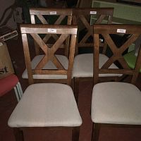 Four wooden dining chairs with X-back design and beige upholstered seats, arranged together showing fronts and seat upholstery.