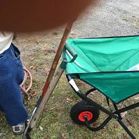 Pitch fork leaning against a green nylon garden wheelbarrow with black frame and red wheels on grass.