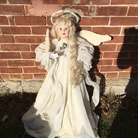 Full frontal view of the porcelain angel figurine standing against a brick wall, showing dress, wings, and halo.