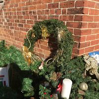 Photo showing green artificial Christmas wreath with gold bow, pine cone ornaments, and additional decorative elements, set against brick wall.