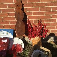 Photo showing rusty metal snowman garden stake, basket with faux cranberry branches and fern leaves, white plates with holly sprig designs, and gift boxes with red snowflake patterns.
