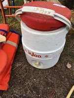 White Igloo water cooler with red lid and spout, sits on ground outdoors