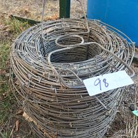 Top side view of a tightly coiled roll of barbed wire sitting on the ground outdoors with a tag labeled '98'.