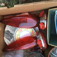 Two red glass hummingbird feeders in a cardboard box, one appears to be blown glass with red flower feeding ports and metal lids